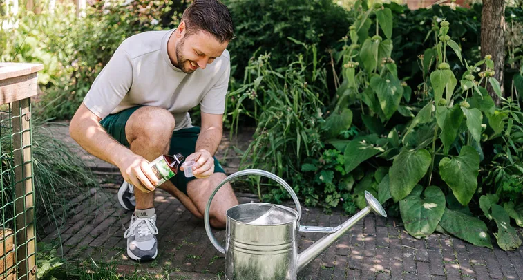 Man giet vloeistof uit een fles in een maatdop naast een metalen gieter, geknield op een stenen pad tussen dichte struiken en tuinplanten. Tekst op schoen: "HUB".