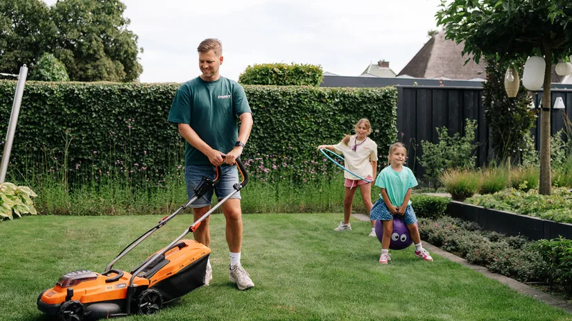 Gazon in de zomer.png Man duwt een oranje grasmaaier over een net gazon terwijl twee meisjes achter hem spelen: één zit op een springbal, de ander zwaait met een hoepel in een besloten tuin.