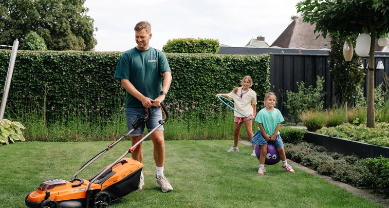 Man duwt een oranje grasmaaier over een net gazon terwijl twee meisjes achter hem spelen: één zit op een springbal, de ander zwaait met een hoepel in een besloten tuin.